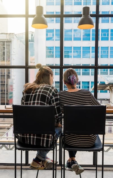 Two colleagues in a brightly lit office, one sitting across from the other. One is attentively listening with a slight smile, while the other is speaking, gesturing with their hands to emphasize their point.