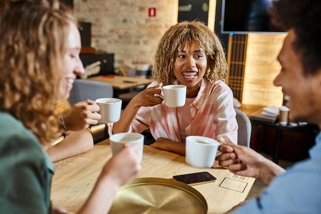 A diverse group of colleagues smiling and laughing together in an office break room, one person holding a cup of tea and others chatting animatedly, surrounded by plants and comfortable seating.