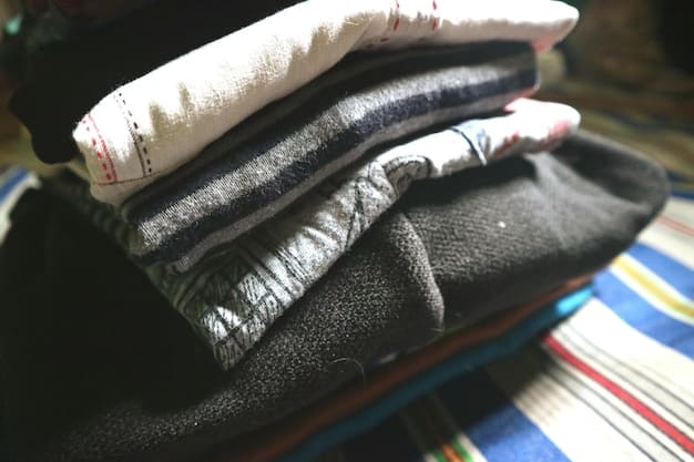 A close-up of a neatly folded pile of clothing, including a lightweight down jacket, a merino wool sweater, and a pair of dark wash jeans. The focus is on the fabric and textures of these versatile travel items.