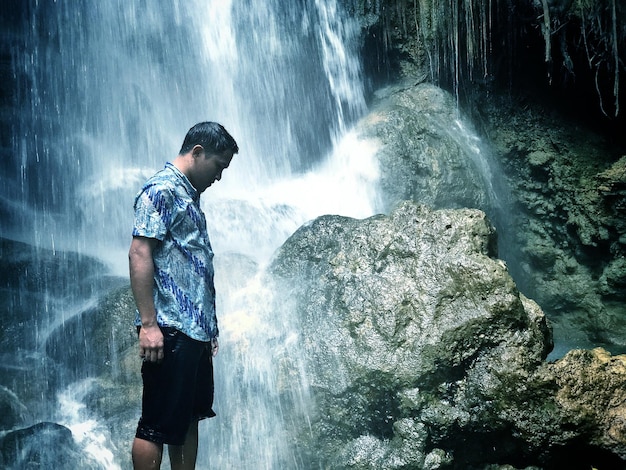 A medium shot of a hiker standing near the base of a waterfall, wearing hiking boots and a backpack, looking up at the cascading water with awe and admiration. The water is crystal clear, and the surrounding rocks are covered in moss.
