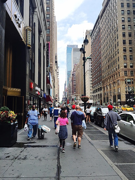 A crowded street corner in New York City. People are rushing by, and there are several funny signs and posters visible in the background. The image captures the frenetic energy and diverse characters of the city.